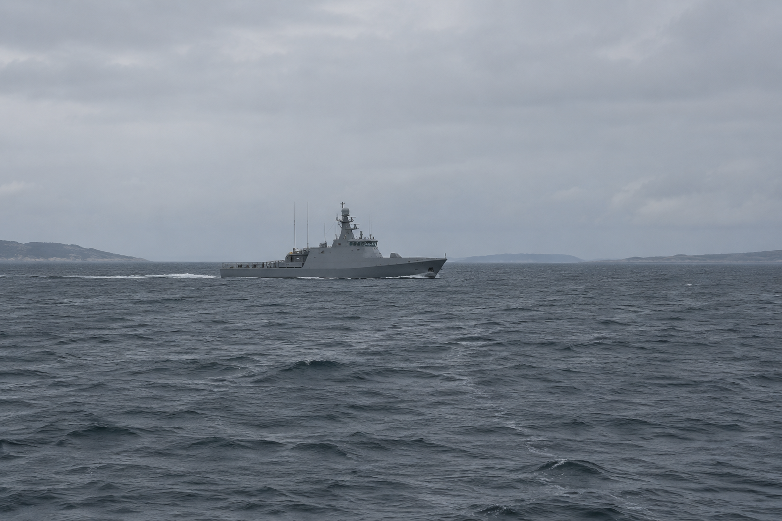 A grey naval patrol vessel under way in open coastal water, low headland on the horizon