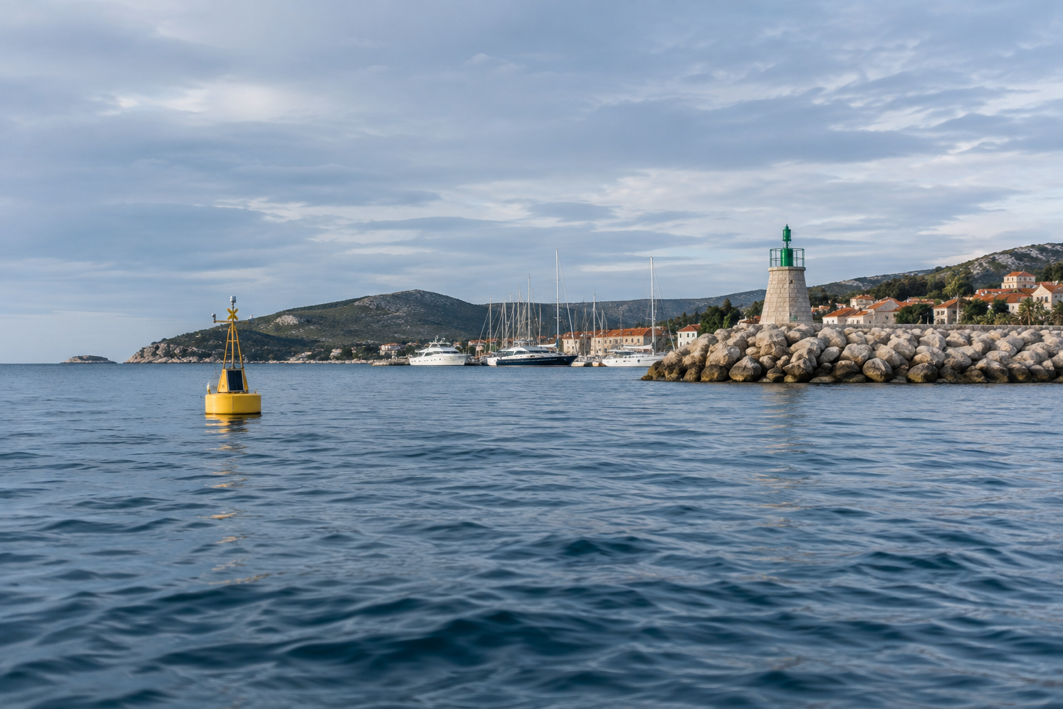 A harbour entrance seen from the water, a yellow monitoring buoy in the foreground and a stone breakwater with a navigation light beyond it