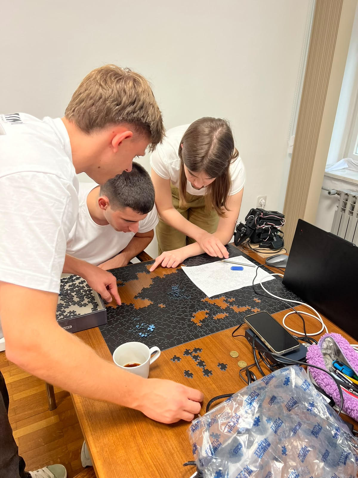 Three students leaning over a table at MARBLE headquarters, assembling a large jigsaw puzzle beside an open laptop and hardware